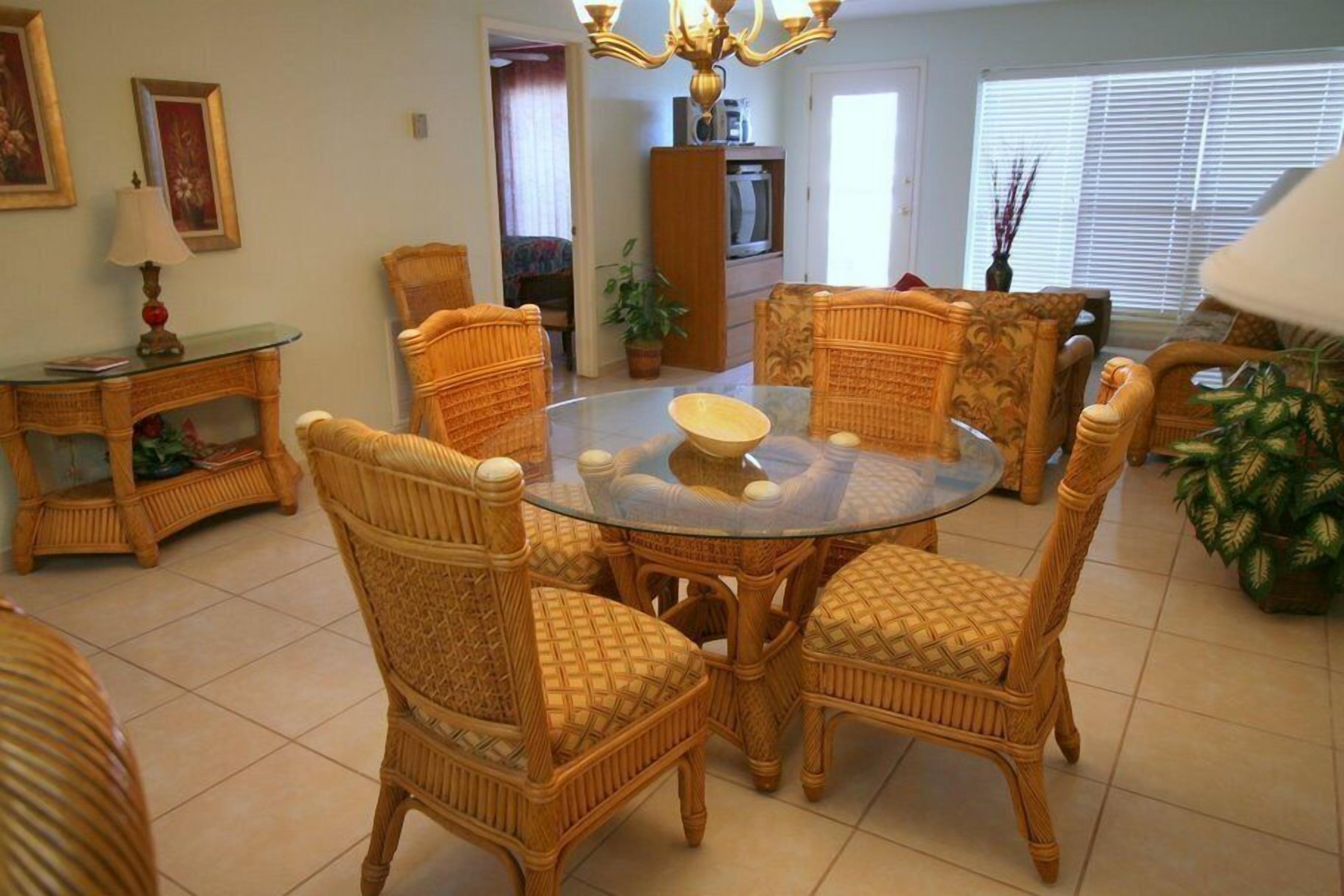 Un comedor con sillas de mimbre y una mesa de vidrio, rodeado de un piso de baldosas y una planta en maceta.