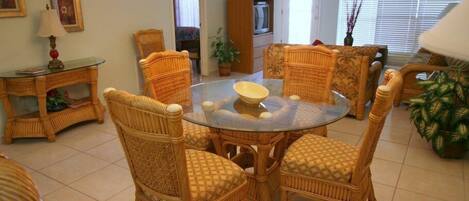 Un comedor con sillas de mimbre y una mesa de vidrio, rodeado de un piso de baldosas y una planta en maceta.
