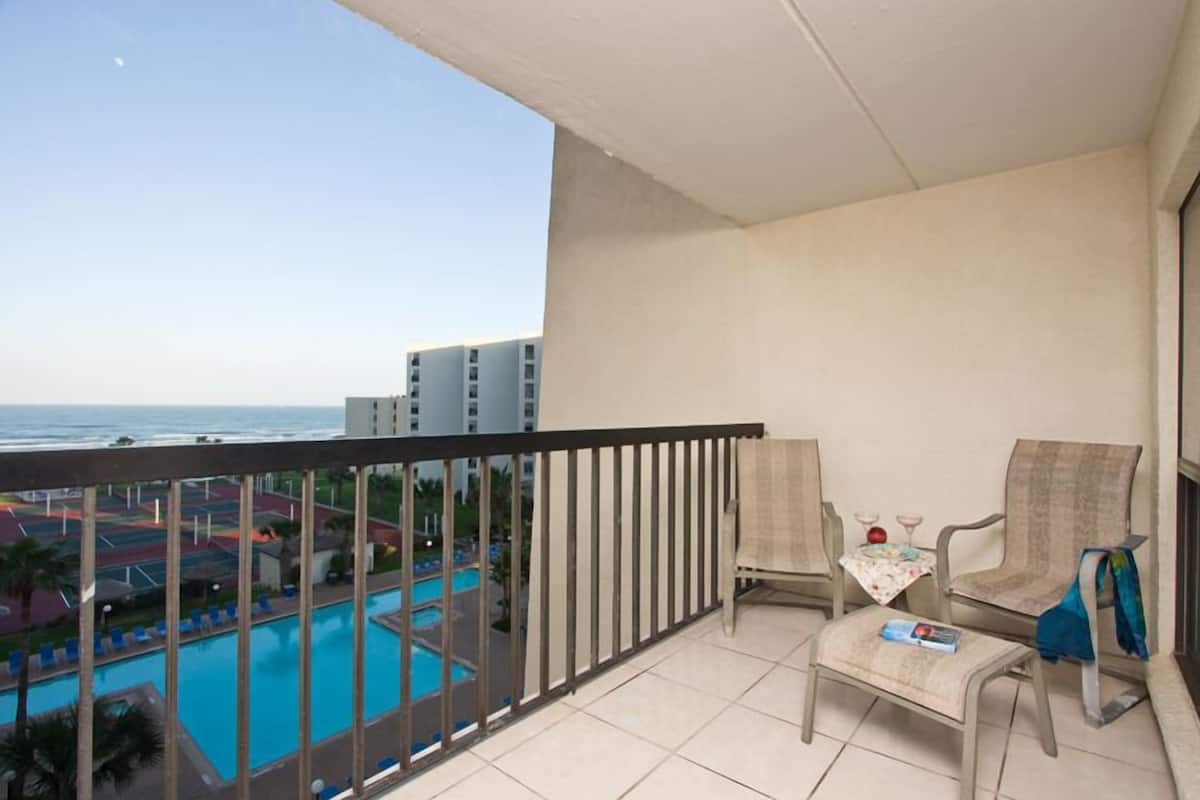 A balcony with a view of a swimming pool and a beach. There are two chairs and a small table with a book and a glass.