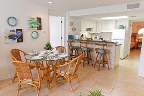 A dining area with a round glass table surrounded by wicker chairs, a kitchen with white cabinets and a central island, and artwork on the walls. - Bahia Mar by Padre Island Rentals (South Padre Island)