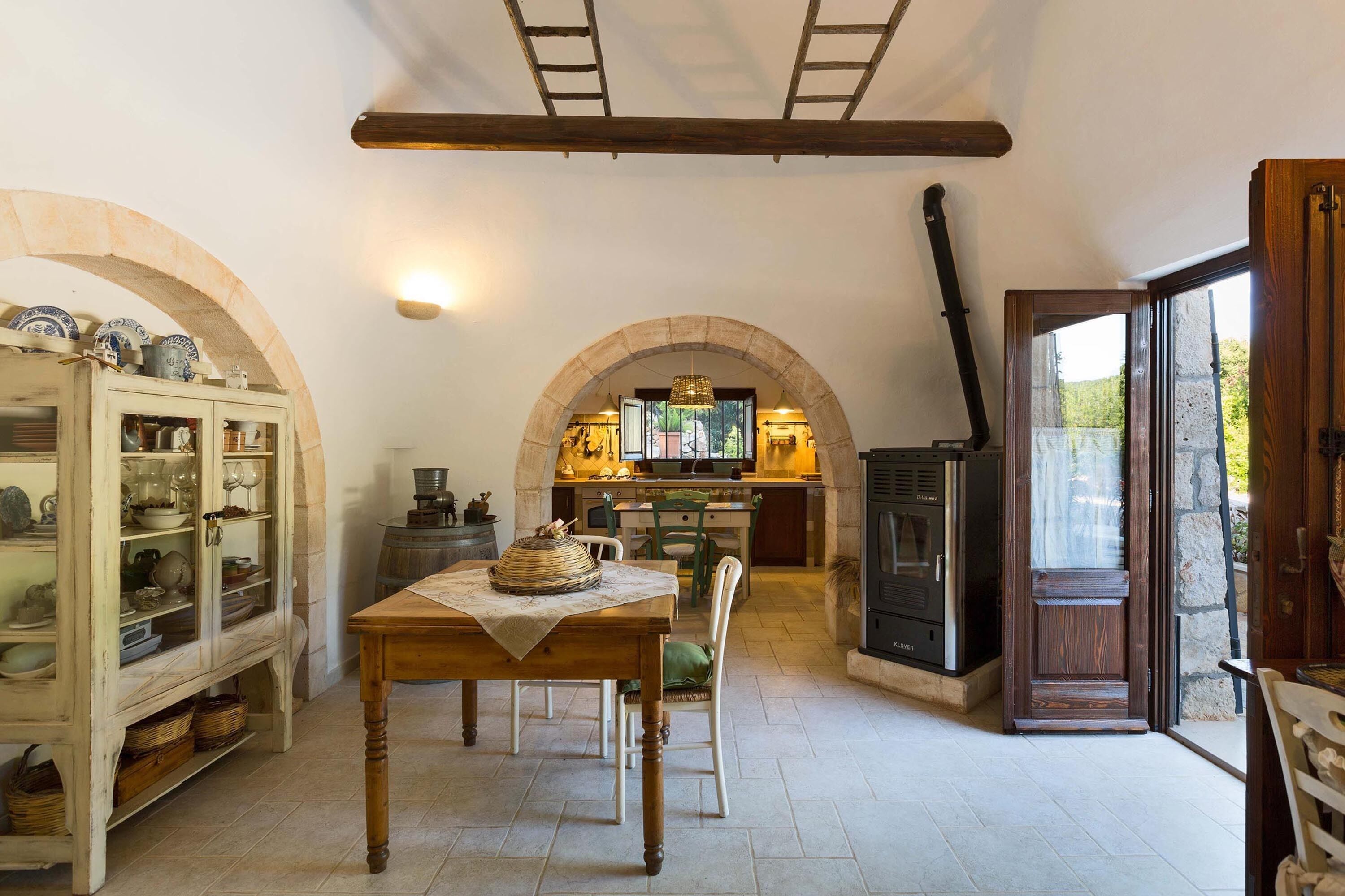 A rustic kitchen with a wooden table, chairs, and an open archway leading to a dining area.