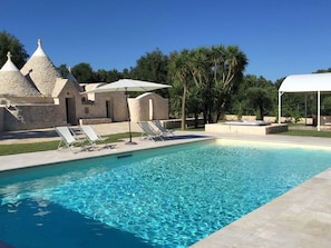 Une piscine à l’eau d’un bleu cristallin, entourée de chaises longues et de parasols, avec des bâtiments en pierre et des palmiers en arrière-plan.