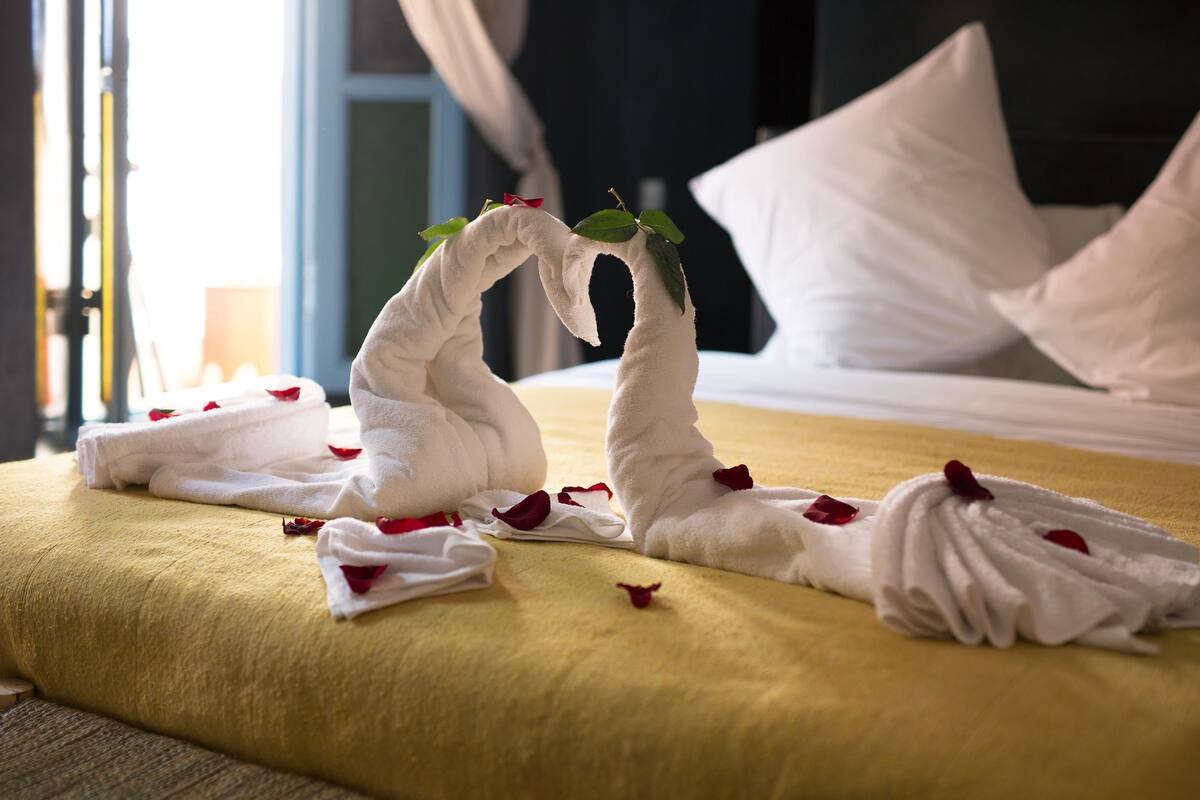 A bed with white towels arranged in a heart shape, adorned with red rose petals and green leaves.