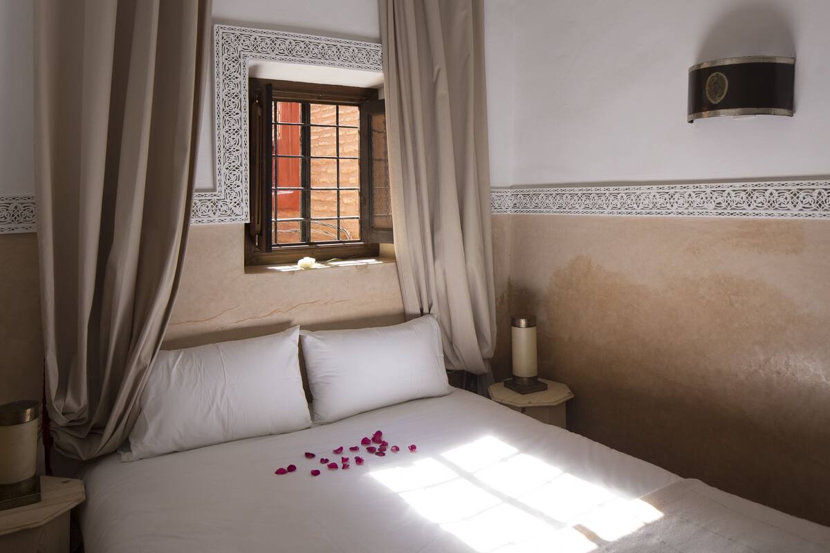 A bed with white linens, a window with a decorative frame, and a wall-mounted light fixture.