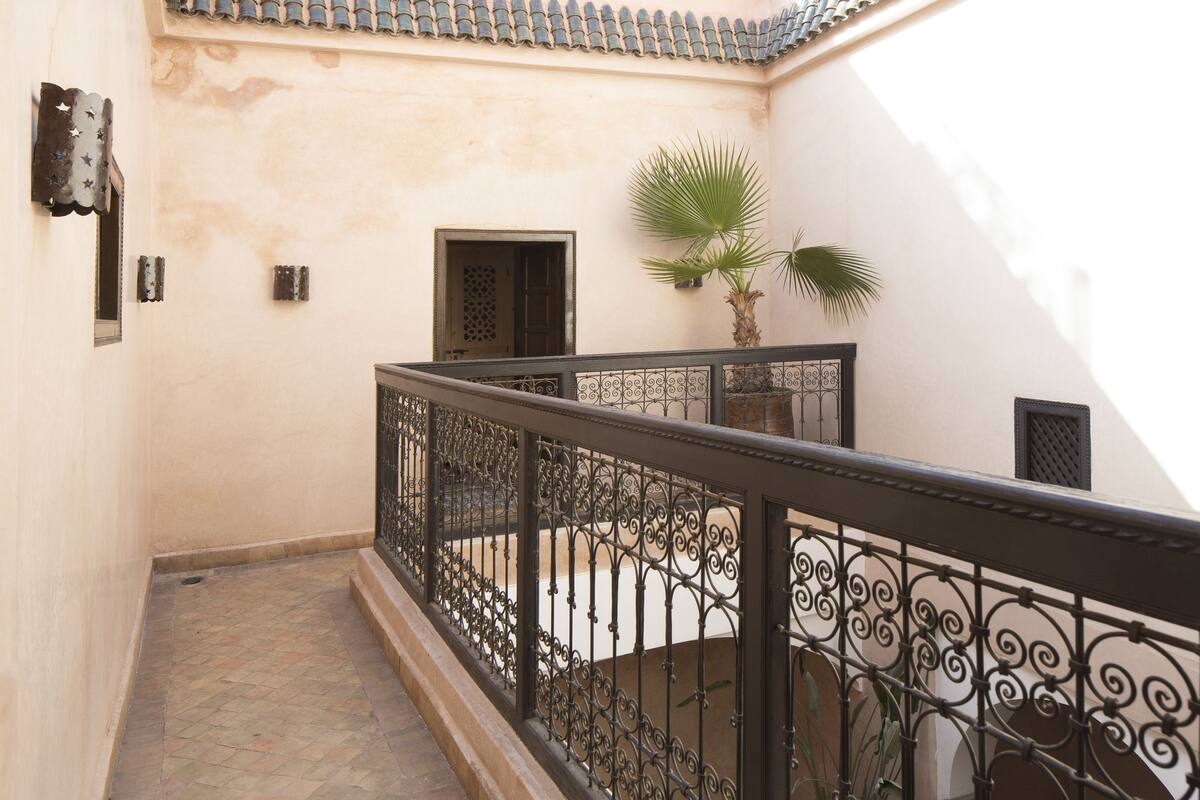 A narrow, tiled corridor with a wrought iron railing, a palm tree, and a door with a decorative grille.