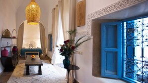 Una habitación con una lámpara colgante de mimbre, una puerta azul con detalles de metal decorativo, una pared blanca con textura, una pequeña mesa redonda con un jarrón de flores y una cama con cabecera azul.