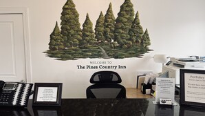 A reception desk at The Pines Country Inn with a welcome sign, a framed 'If no one is in the office' sign, a phone, and a pet policy sign. - The Pines Country Inn (Owosso)