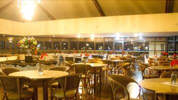 A spacious indoor dining area with round tables and wicker chairs, featuring a large window with a view of the city at night.