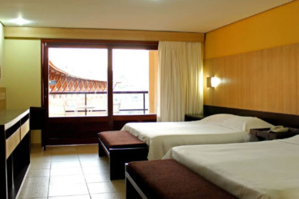 A hotel room with two beds, a desk, a bench, a large window with a view, and a curtain.