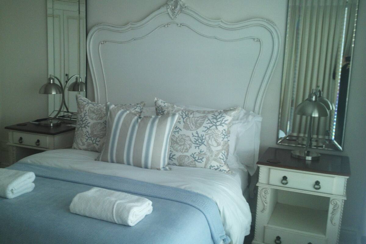 A neatly made bed with a blue bedspread and white pillows, a bedside table with a lamp, and a mirror on the wall.