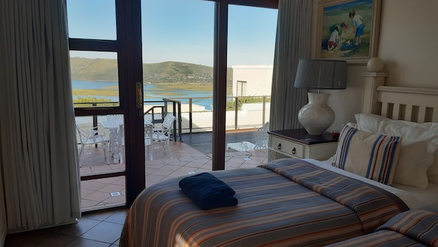 Een hotelkamer met een bed, een nachtkastje, een lamp en een schilderij. Er is een balkon met een tafel en stoelen, en uitzicht op zee en heuvels.