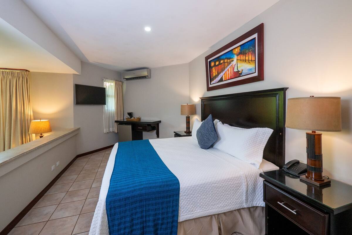 Une chambre d’hôtel comprenant un lit, une table de chevet, une lampe, un bureau, une chaise et un tableau au mur.