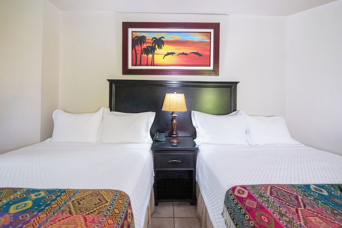 Habitación de hotel con dos camas, una mesita de noche, una lámpara, un cuadro con una imagen de un atardecer y una colcha estampada.