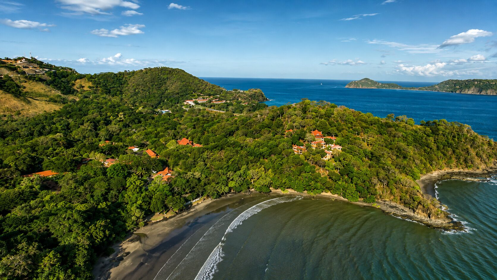 An aerial view of a coastal area with a beach, dense greenery, and buildings with red roofs.