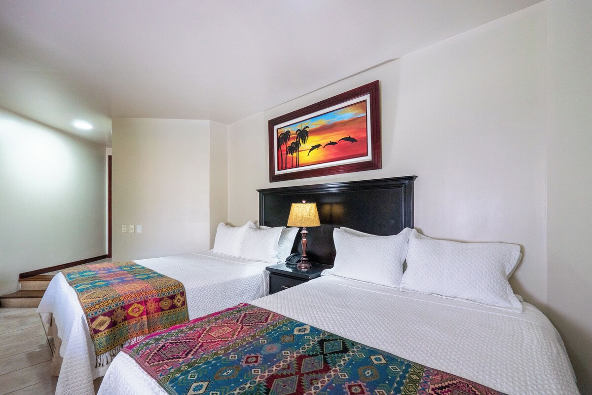 Une chambre d’hôtel avec deux lits, une table de chevet avec une lampe, un tableau représentant un coucher de soleil et un tapis à motifs.