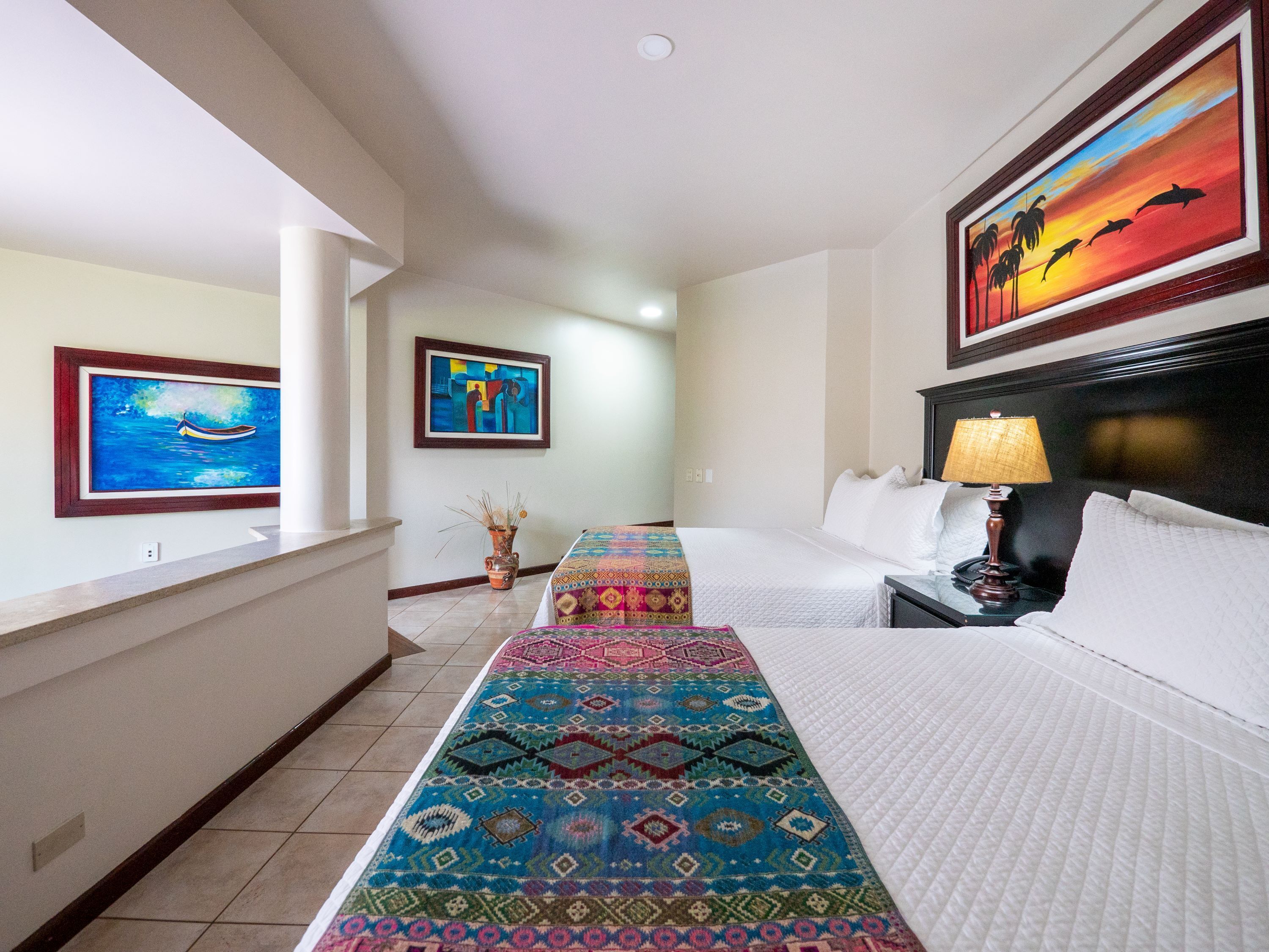 Une chambre d’hôtel avec un lit, une table de chevet, une lampe, un tapis coloré et deux œuvres d’art encadrées accrochées au mur.