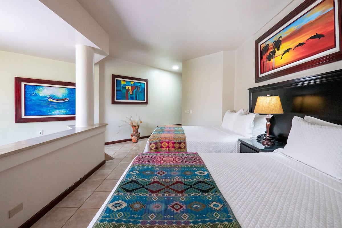 Une chambre d’hôtel avec un lit, une table de chevet, une lampe, un tapis coloré et deux œuvres d’art encadrées accrochées au mur.