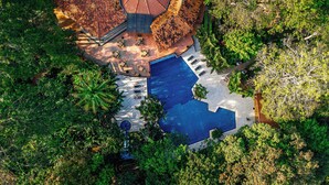 Vue aérienne d’un complexe hôtelier doté d’une grande piscine, entouré d’une végétation luxuriante et comprenant un espace de détente abrité.