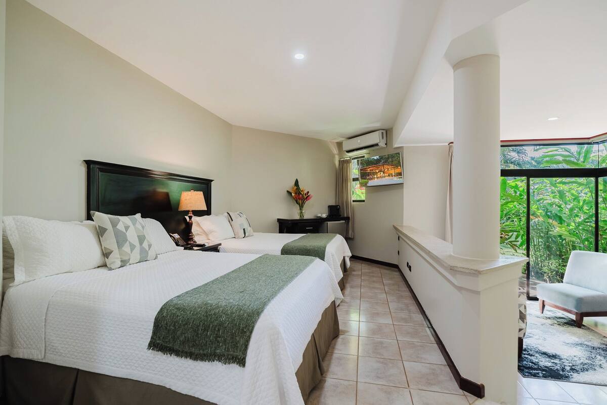 Une chambre d’hôtel avec deux lits, un bureau, une chaise et un balcon donnant sur un espace verdoyant.