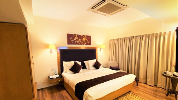 A hotel room with a large bed, a nightstand, a lamp, a window with curtains, and a ceiling vent. - RnB Banjara Hills (Hyderabad)