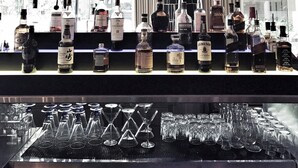 Zona de bar con bombillas colgantes, una variedad de botellas de licor y vasos vacíos sobre la barra.