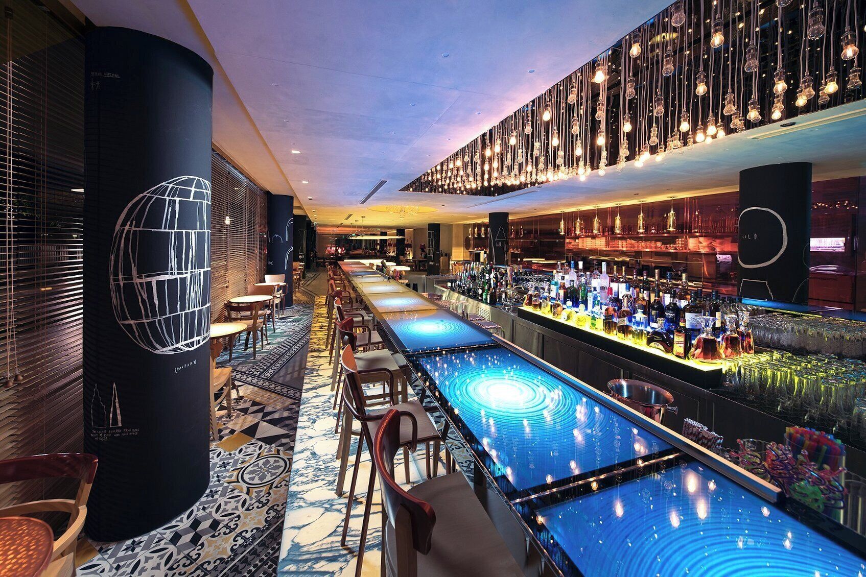 A modern bar area with a long counter, multiple seating options, and a variety of bottles and glasses.