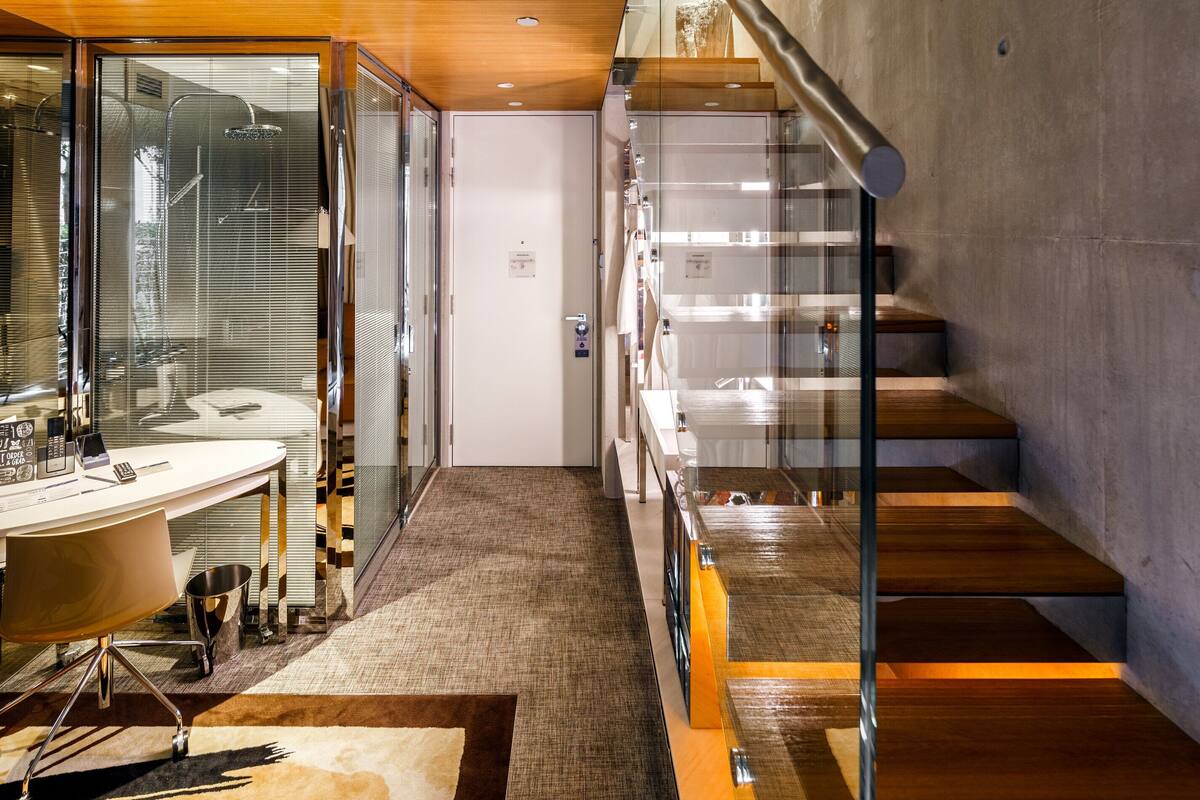 Een moderne hotelkamer met een bureau met glazen wanden, een bed met houten hoofdeinde, en een trap met glazen leuningen.