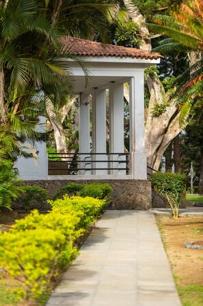 Un pavillon couvert, doté de colonnes en pierre et d’un toit en tuiles, entouré d’une végétation luxuriante et bordé d’un chemin pavé.