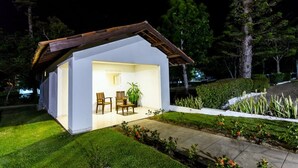 Una casa pequeña y moderna con fachada blanca y techo de madera. En su interior, hay una zona de estar con dos sillas y una mesa, además de una planta en maceta. La casa está rodeada de un césped cuidado y un sendero.