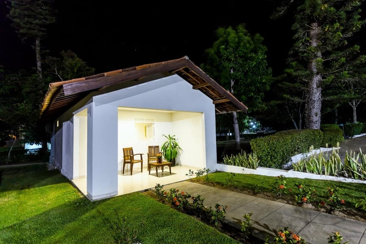 Il s’agit d’une petite maison moderne, à l’extérieur blanc et au toit en bois. À l’intérieur, on trouve un coin salon avec deux chaises et une table, ainsi qu’une plante en pot. La maison est entourée d’une pelouse bien entretenue et d’un chemin.