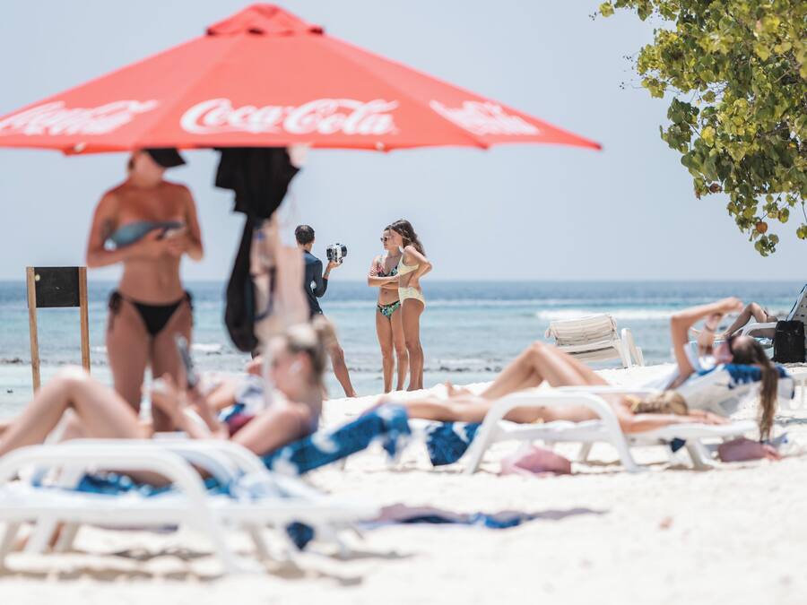 Escena de playa con gente tomando el sol bajo una sombrilla roja, algunos recostados en sillas de salón, y un camarógrafo grabando la escena.