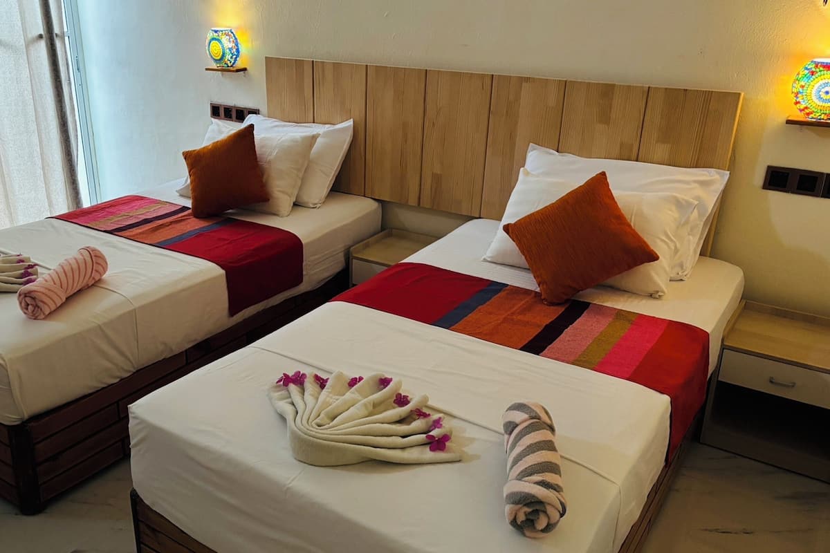 A hotel room with two beds, each with a wooden headboard, white bedding, and a red and orange striped blanket. There are towels and a small table between the beds.