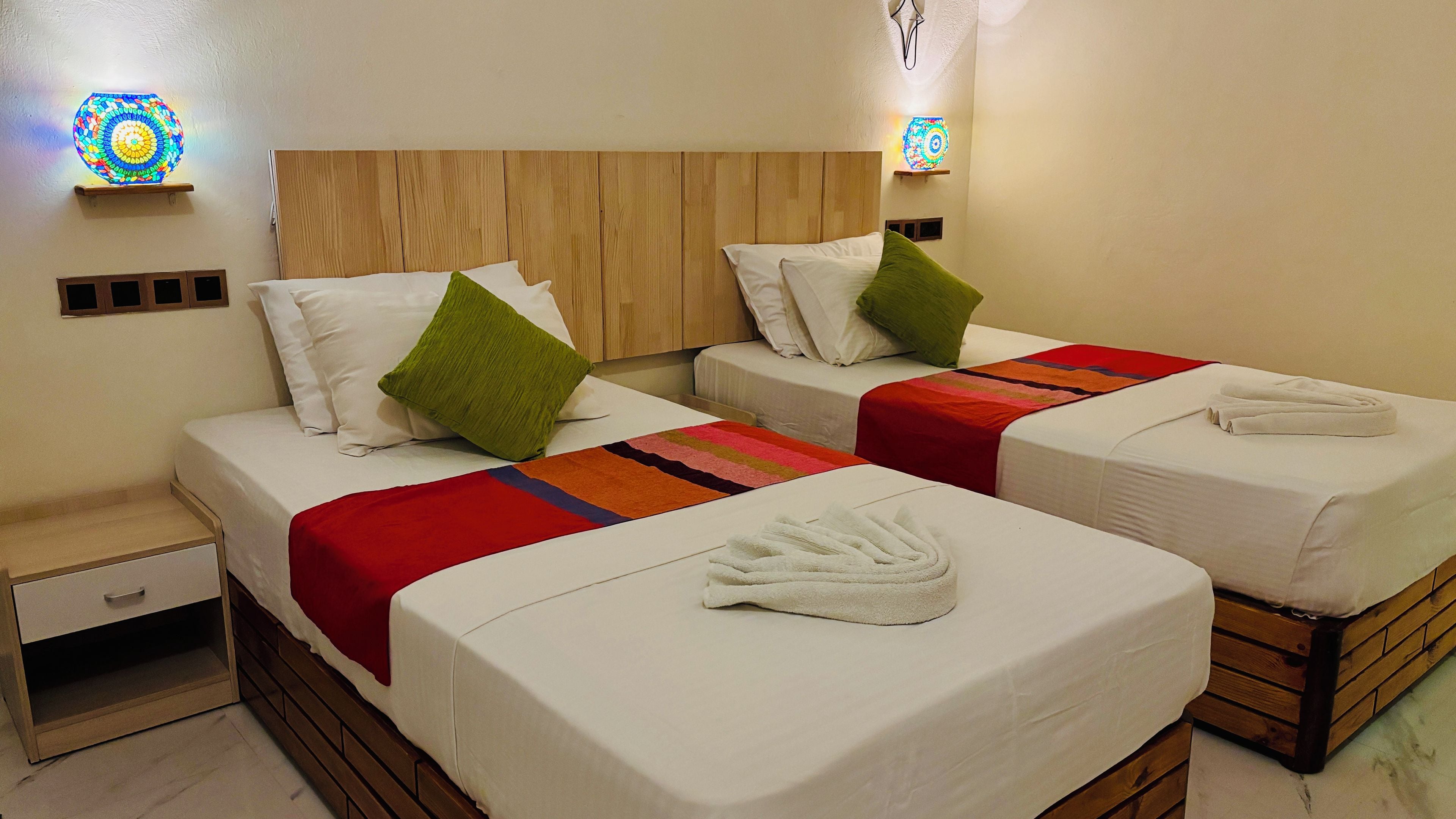 A hotel room with two beds, each with white linens, green pillows, and a red blanket. There are wall-mounted lights and a wooden headboard.