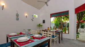 Un comedor con mesas dispuestas para cuatro, sillas blancas con cojines azules y un ventanal con marco rojo que da a un jardín.