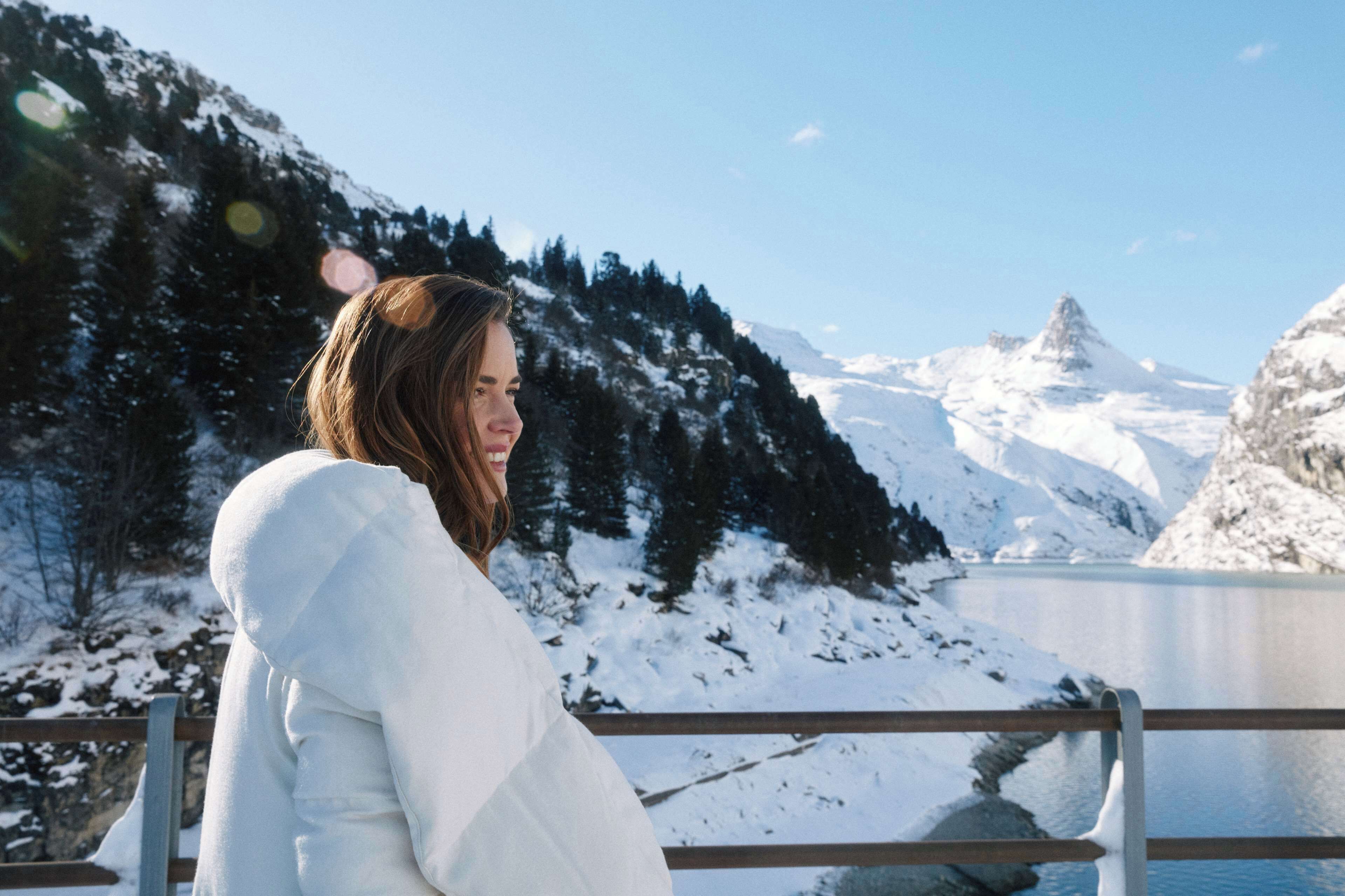 A person in a white coat standing by a railing with a snowy mountain and lake in the background.