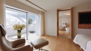 A modern hotel room with a large sliding glass door leading to a balcony with a mountain view. Inside, there is a bed, a seating area with a chair and ottoman, and a bathroom with a bathtub and a large mirror. - 7132 Hotel (Vals)