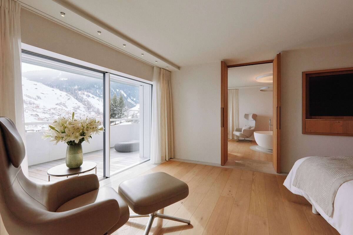 Une chambre d’hôtel moderne dotée d’une grande porte-fenêtre coulissante donnant sur un balcon avec vue sur les montagnes. L’intérieur comprend un lit, un coin salon avec un fauteuil et un pouf, ainsi qu’une salle de bain avec une baignoire et un grand miroir.