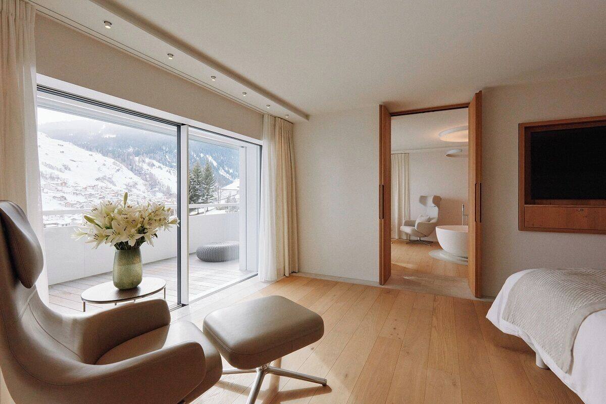 Une chambre d’hôtel moderne avec une grande fenêtre, un lit, une chaise et une salle de bain visible à travers une porte ouverte.