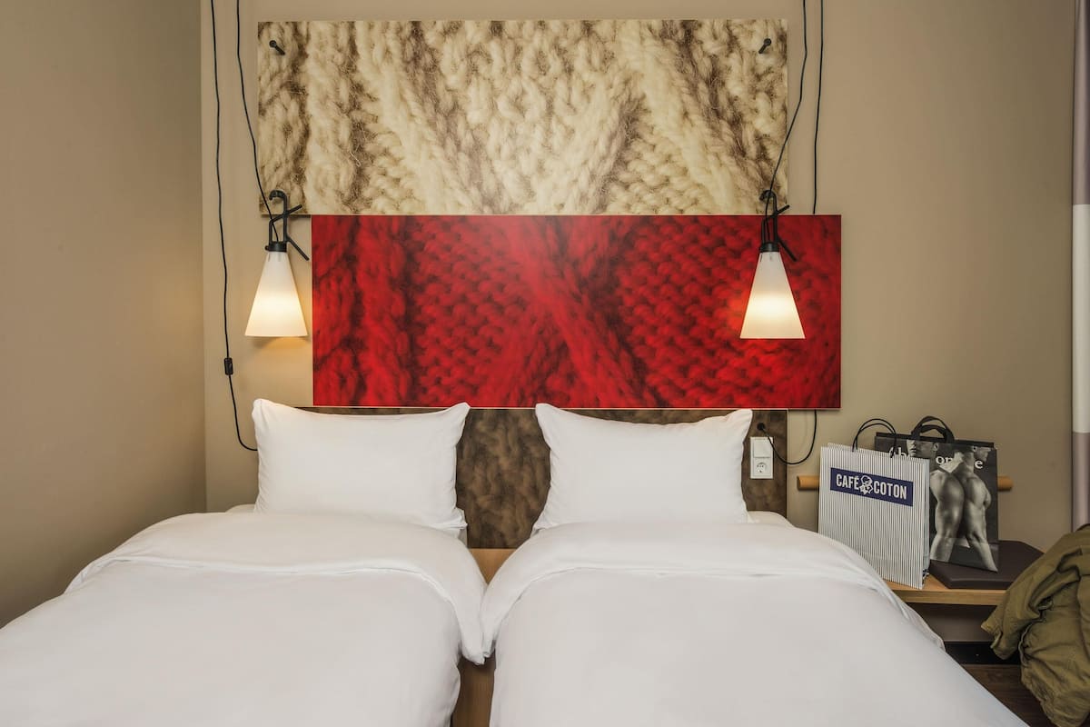 Habitación de hotel con dos camas, cabecera con diseño texturizado y mesita de noche con lámpara y bolso.