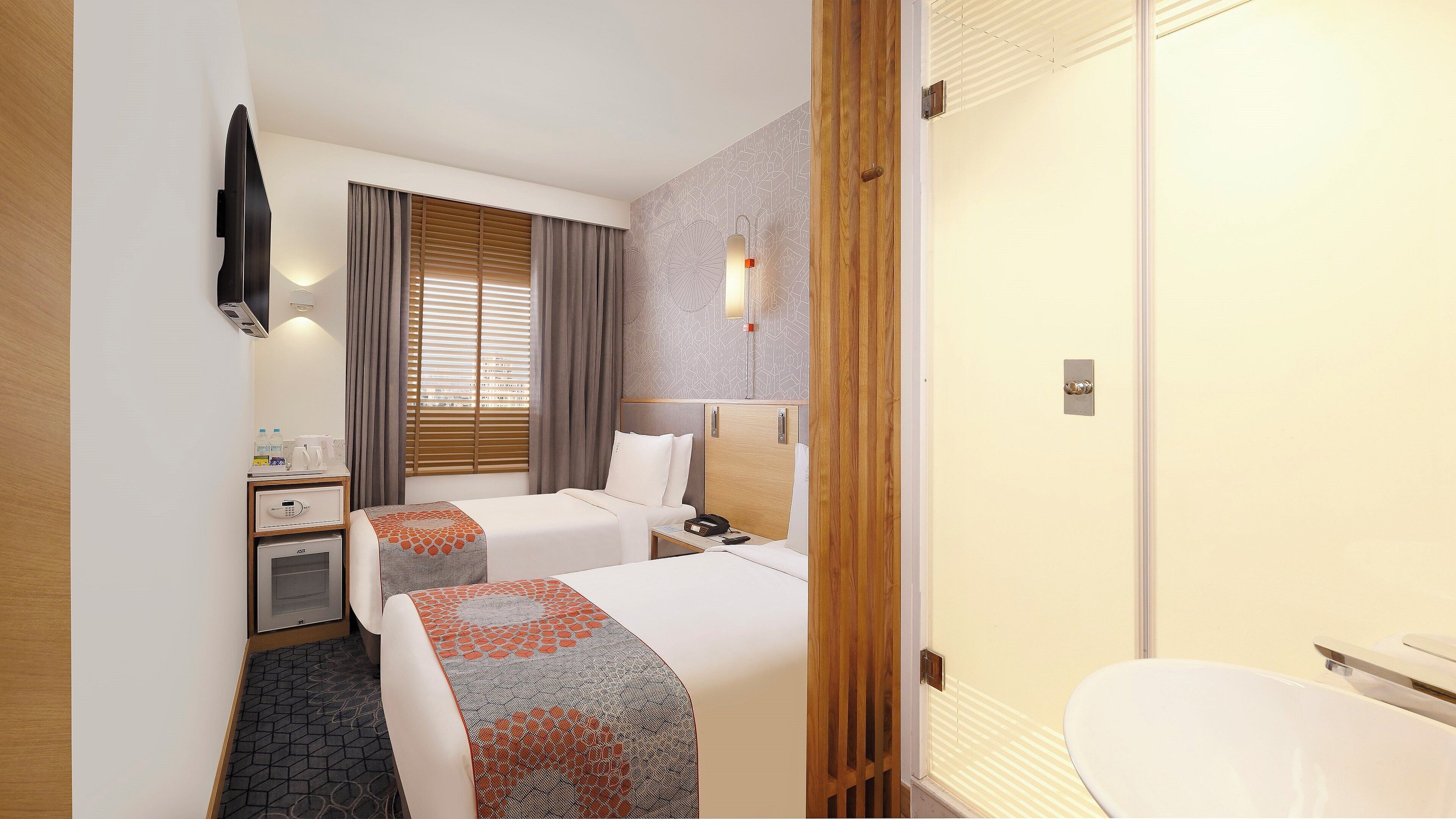Habitación de hotel con dos camas, escritorio, minibar, baño con inodoro y lavabo, y ventana con persianas.