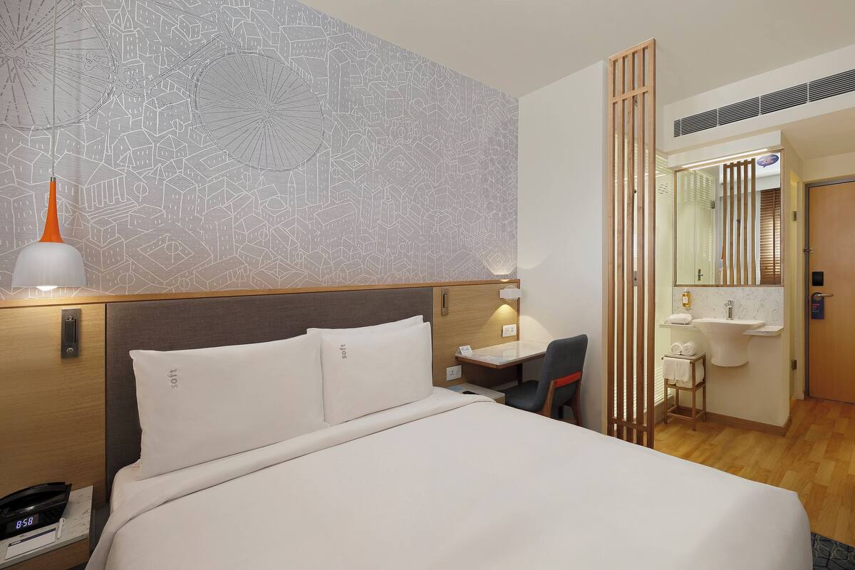 Ett hotellrum med en säng, ett skrivbord, en stol, ett litet bord med en vattenkokare och en kaffebryggare, en spegel och ett badrum som syns genom en avskärmning.