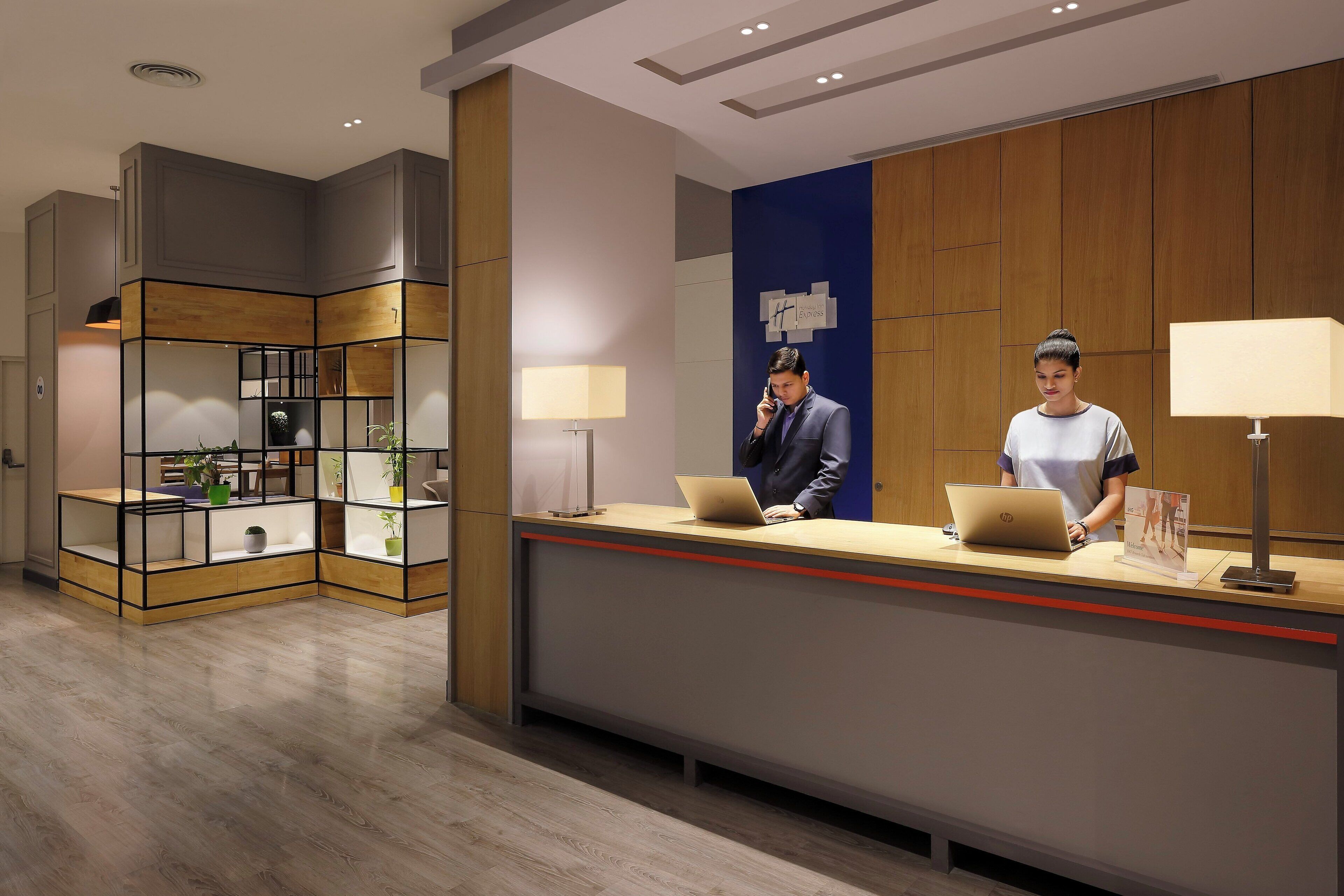A hotel reception desk with two employees, a blue wall with a logo, and a modern shelving unit with plants.