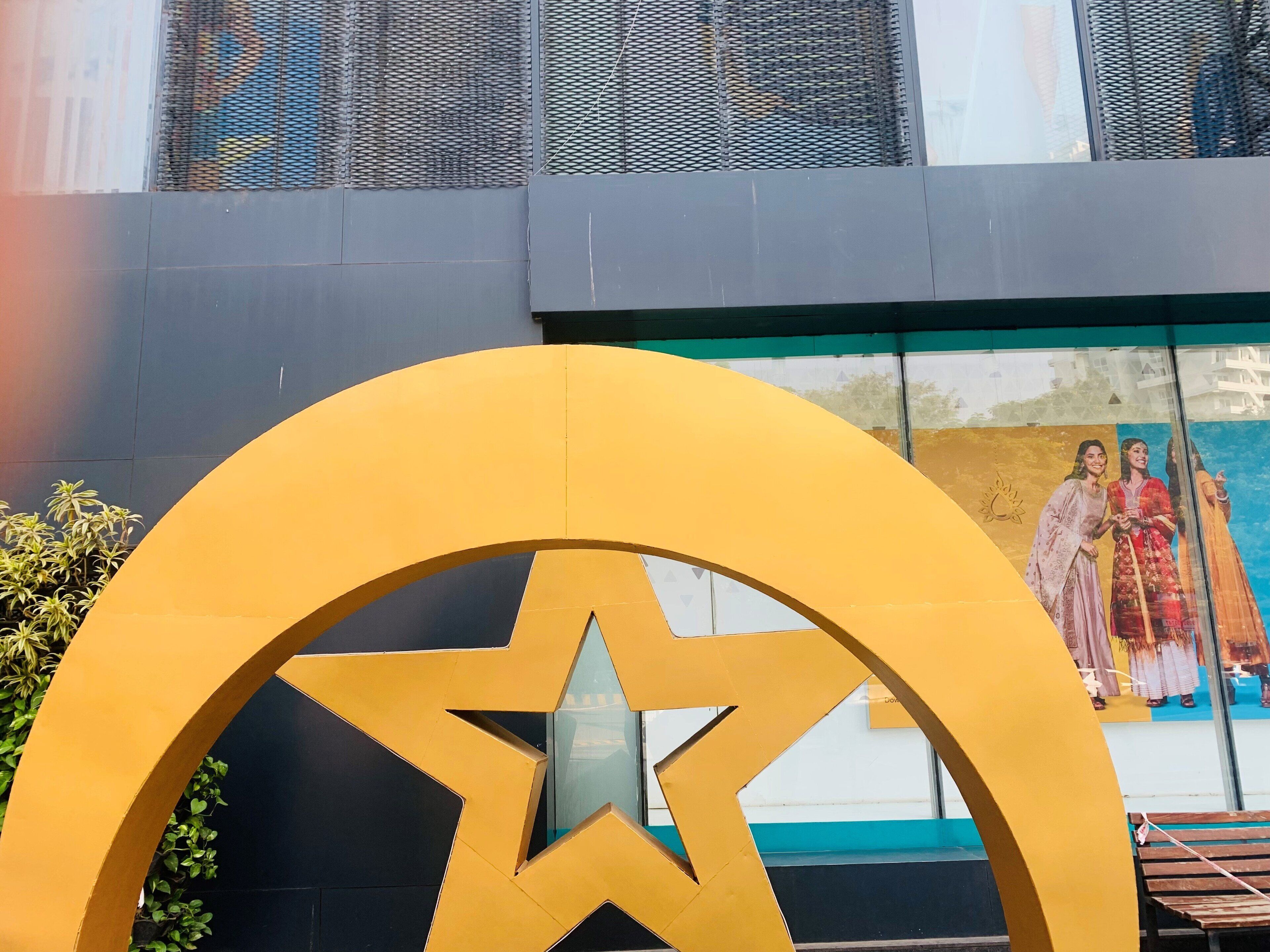 Una gran estructura en forma de estrella amarilla con un ventanal de vidrio en el centro, frente a la fachada de un edificio con una ventana de malla y un mural que representa a dos figuras.