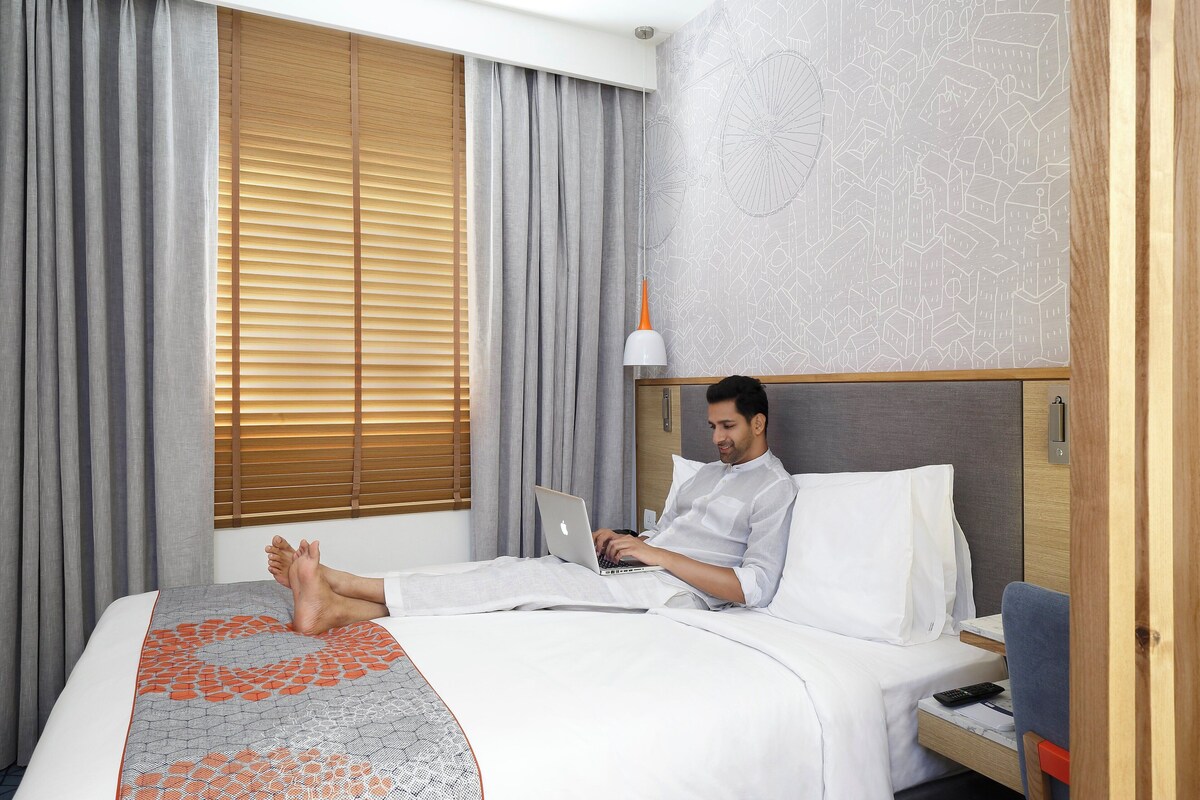 Une chambre d’hôtel avec un lit, un ordinateur portable, une table de chevet et une fenêtre avec des stores.