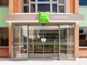 A hotel entrance with a glass door, a green sign above the door, and a sign indicating 'London Heathrow Airport' to the right.