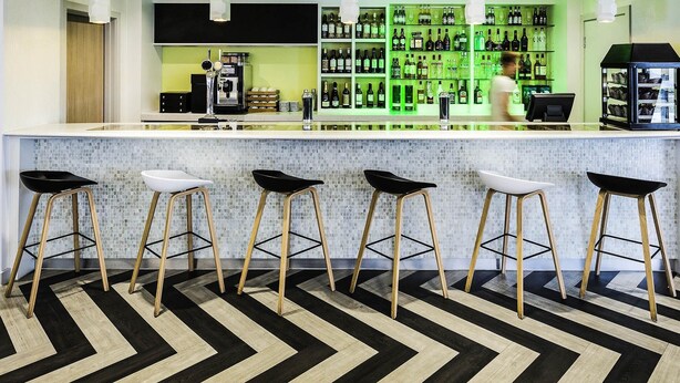 A modern bar area with a bar counter, stools, and a green-lit shelving unit displaying bottles. - Ibis Styles London Heathrow (West Drayton)