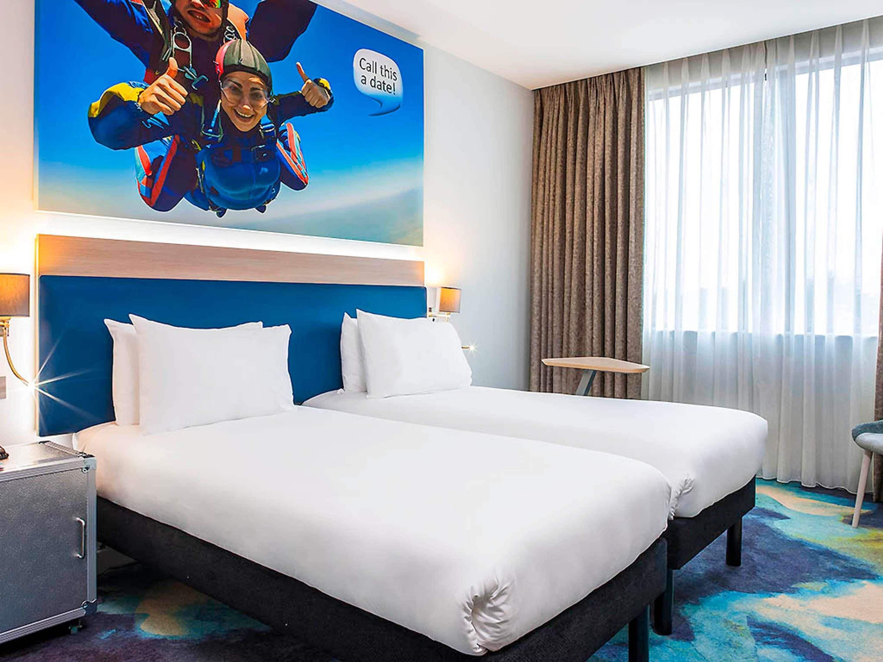 A hotel room with a large bed, a headboard, a nightstand, a lamp, a small table, and a window with curtains.