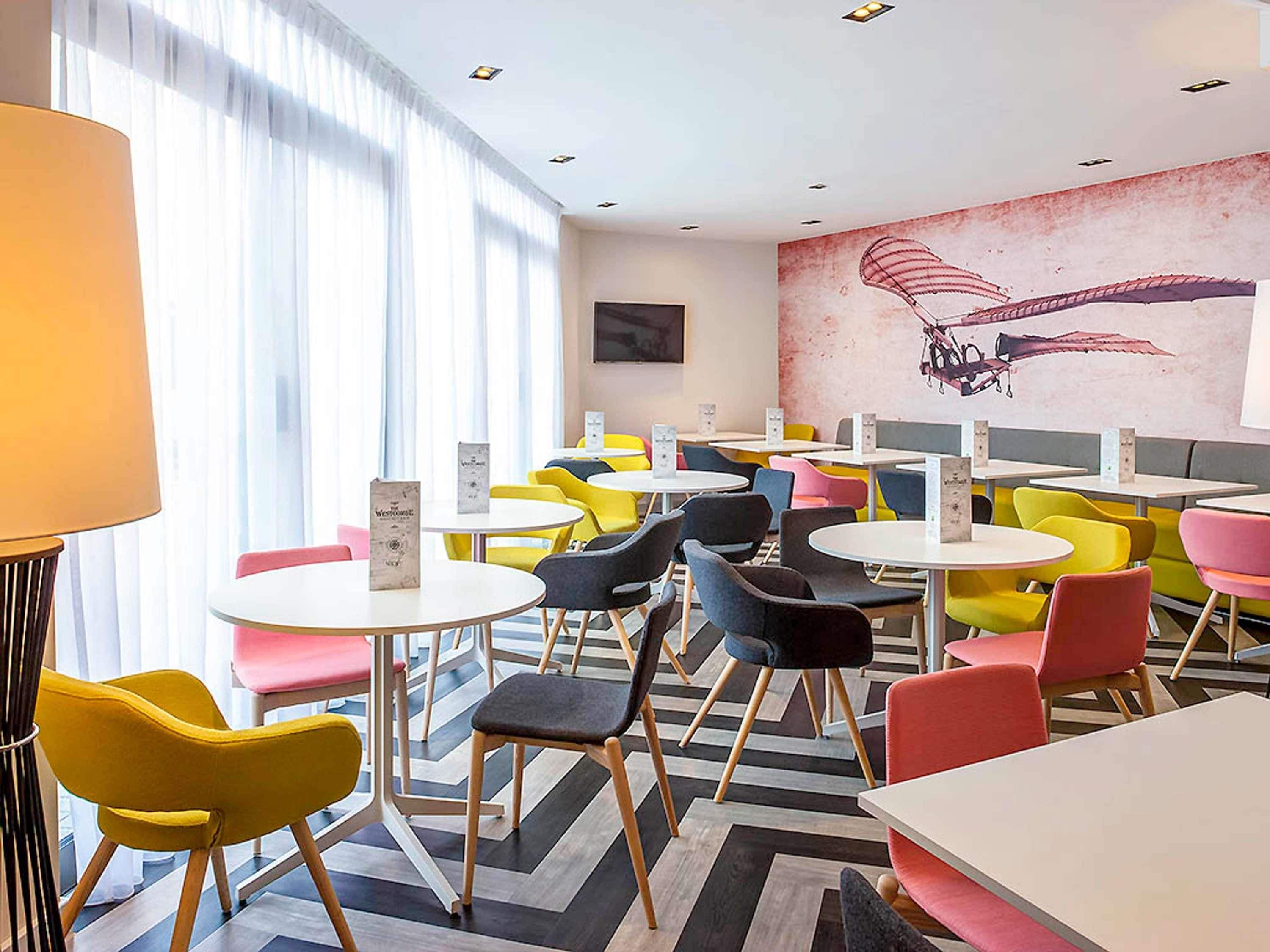 A modern cafe with round tables and colorful chairs, a mural on the wall, and a large window with sheer curtains.