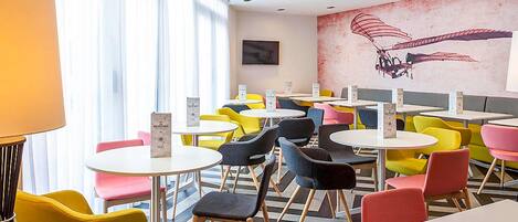 Un café moderno con mesas redondas y sillas coloridas, un mural en la pared y un amplio ventanal con cortinas translúcidas.