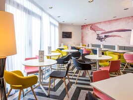 A modern cafe with round tables and colorful chairs, a mural on the wall, and a large window with sheer curtains.